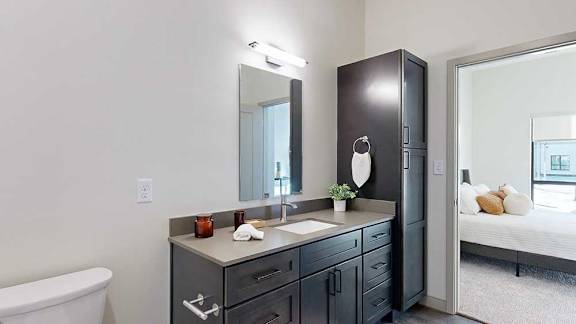 A bathroom with a quartz vanity, dark wooden cabinets, a tall storage cabinet, and a door open to a bedroom.