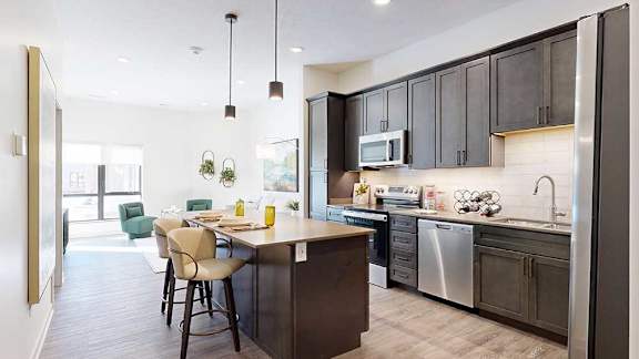 A kitchen with dark wooden cabinets and a large island, in front of a spacious living room with floor-to-ceiling windows.