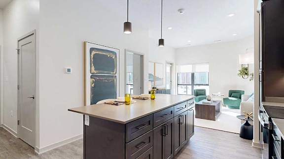A kitchen with dark wooden cabinets and a large island, in front of a spacious living room with floor-to-ceiling windows.