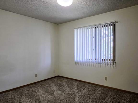 A carpeted bedroom with a window covered by white vertical blinds.
