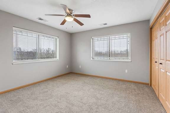 Spacious apartment bedroom with neutral carpet, gray walls, a ceiling fan, two windows with blinds providing natural light, and double bi-fold closet doors.