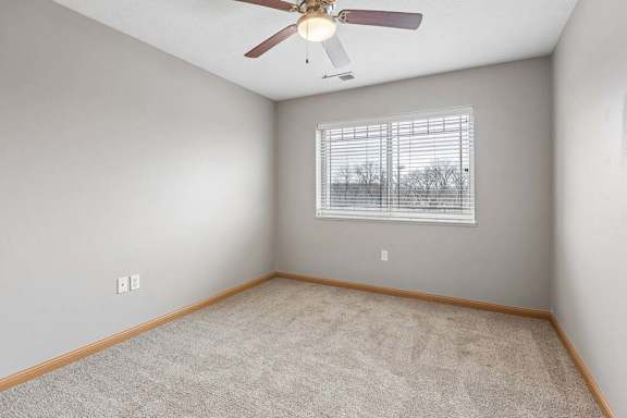 Bright apartment bedroom with neutral carpet, gray walls, a ceiling fan, and a large window with blinds providing natural light.