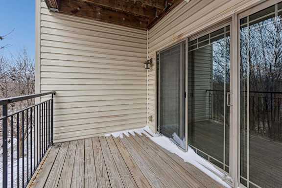 Private apartment balcony with wood decking, metal railing, and sliding glass doors, overlooking a wooded view with trees in the background.