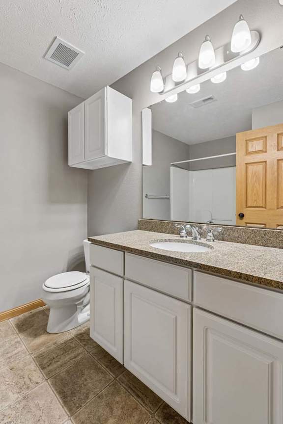 Apartment bathroom with a single-sink vanity, wood cabinetry, laminate countertop, large mirror, modern vanity lighting, and a toilet.