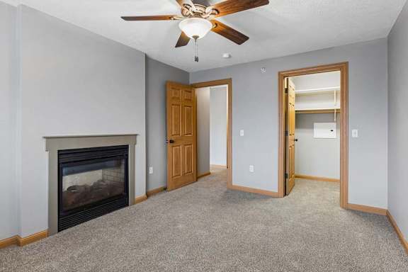 Apartment living room with neutral carpet, a corner fireplace, ceiling fan, and an open doorway leading to a laundry area with shelving.