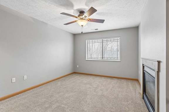 Bright apartment living room with neutral carpet, a corner fireplace, ceiling fan, and a large window with blinds that provides natural light.
