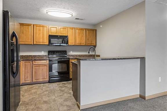Open-concept apartment kitchen with wood cabinets, black appliances, laminate countertops, and a breakfast bar connecting to the adjacent living area.