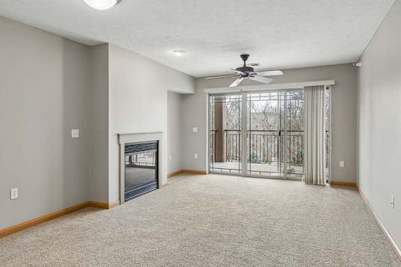 Spacious apartment living room with neutral carpet, a corner fireplace, and sliding glass doors that open to a private balcony with wooded views.