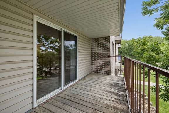 Covered private balcony with wood decking, a sliding glass door, metal railing, and leafy green views.