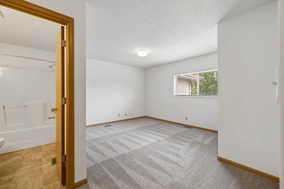 Bright, empty apartment bedroom with fresh carpeting, a large window, neutral walls, wood trim, and an attached bathroom.