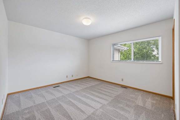 Spacious carpeted bedroom with a wide window overlooking leafy trees, white walls, and ample open floor space.
