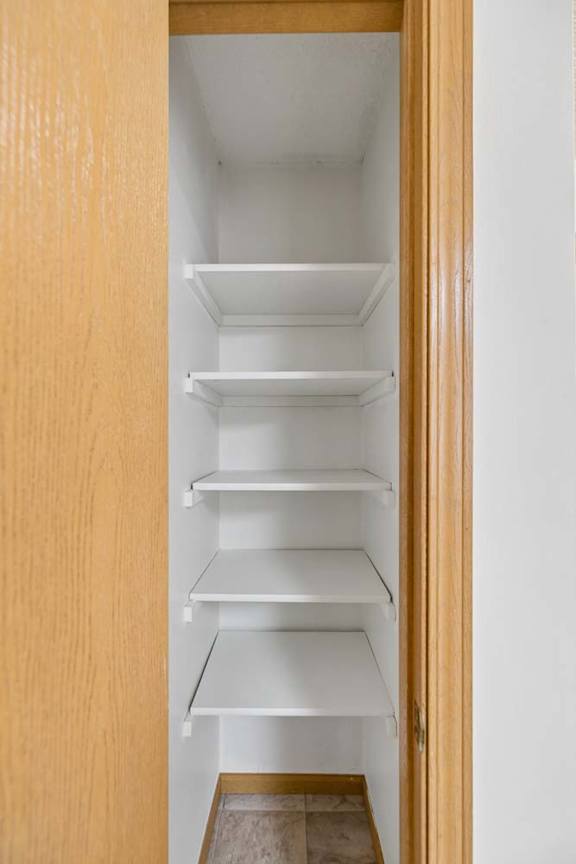 Tall storage closet with five white shelves, a wood door and frame, and tile flooring for organized apartment storage.