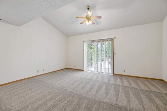 Carpeted living room with a vaulted ceiling, ceiling fan, and sliding glass door opening to a private outdoor area.