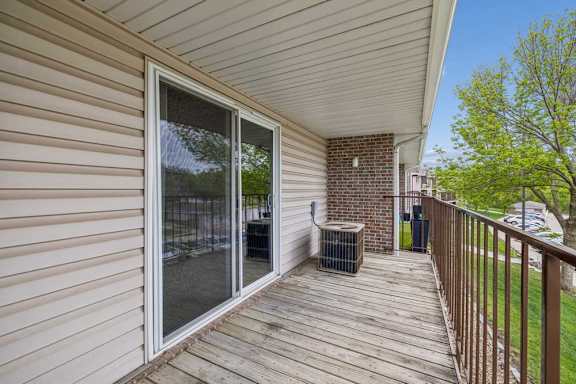 Private apartment balcony with wood decking, sliding glass door, metal railing, and views of mature trees and the surrounding community.