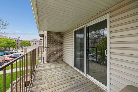 Spacious apartment balcony with wood decking, a sliding glass door, metal railing, and views of mature trees, nearby buildings, and community parking.