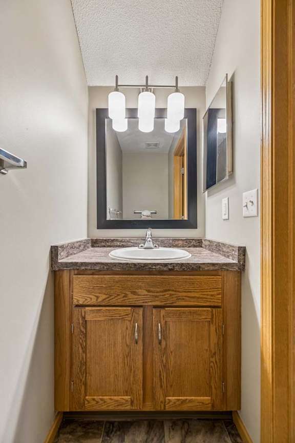 Compact apartment bathroom vanity with a wood-tone cabinet, laminate countertop, framed mirror, modern light fixture, and towel bar in a neutral interior.