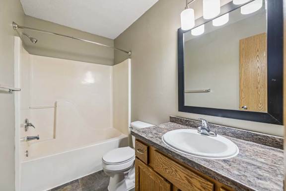 Apartment bathroom with a single-sink vanity, wood-tone cabinet, framed mirror, modern light fixture, toilet, and a tub/shower combination with built-in shelving.