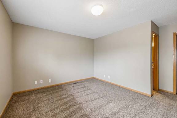 Empty apartment bedroom with neutral carpet, beige walls, wood trim, and an open doorway leading to the adjoining hallway.