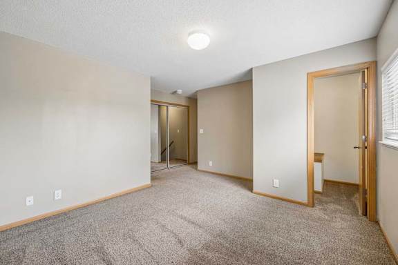 Bright, empty apartment bedroom with neutral carpet, beige walls, wood-trimmed doors, a window for natural light, and an open doorway leading to the closet area.
