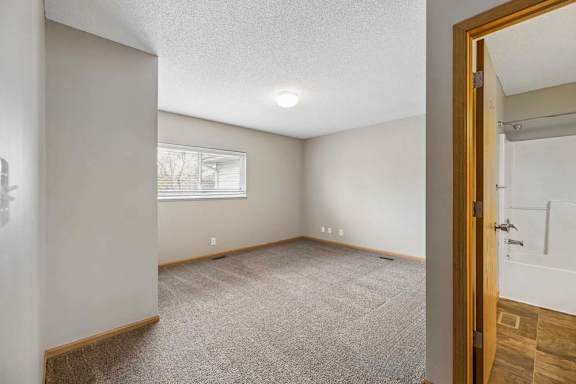 Bright apartment bedroom with neutral carpet, a window for natural light, and an adjoining bathroom with a single vanity and tub/shower visible through the open doorway.