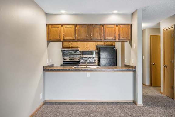 Open apartment kitchen with a breakfast bar, wood-tone cabinets, black appliances, laminate countertops, and a view into the adjacent living area and hallway.