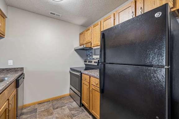 Galley-style apartment kitchen with wood-tone cabinets, black refrigerator and oven, laminate countertops, and durable flooring in a functional layout.