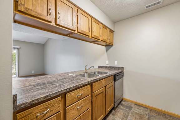 Open apartment kitchen with wood-tone cabinets, laminate countertops, a double-basin sink, dishwasher, overhead storage, and a breakfast bar overlooking the living room.