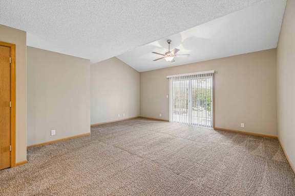 Spacious, empty apartment living room with vaulted ceilings, neutral carpet, a ceiling fan, and a large sliding glass door with vertical blinds that fills the space with natural light.