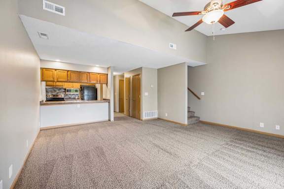Bright, open-concept apartment living room with neutral carpet, a ceiling fan, breakfast bar overlooking the kitchen, wood-tone cabinets, and a staircase leading to the upper level.
