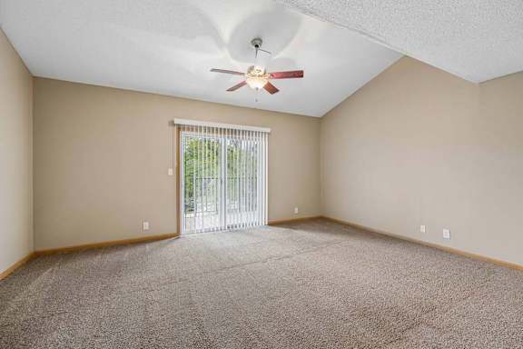 Bright, empty apartment living room with vaulted ceilings, neutral carpet, ceiling fan, and a large sliding glass door with vertical blinds that lets in natural light.