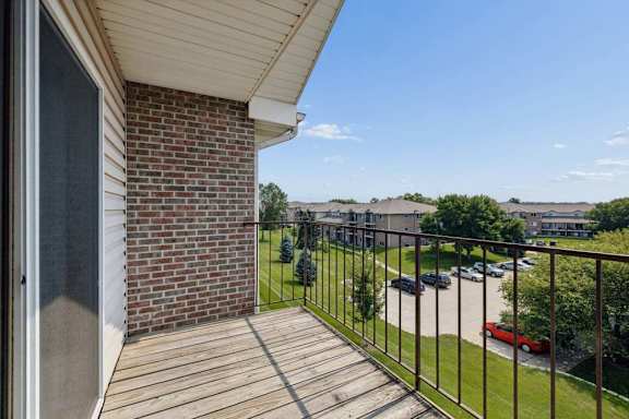 Upper-level balcony with wood decking and metal railing overlooking landscaped community grounds and nearby apartment buildings.
