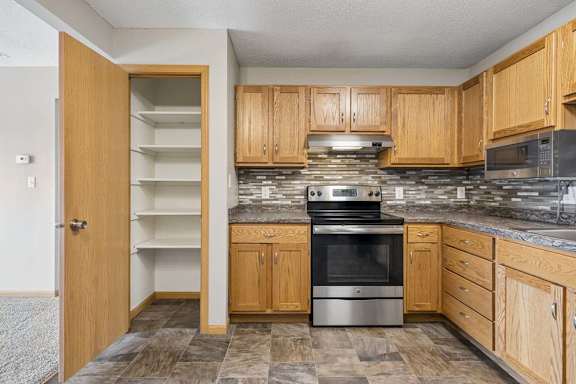 Kitchen with oak cabinets, stone-look counters, mosaic backsplash, stainless oven and microwave, plus a tall shelved pantry.