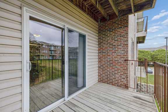 Covered apartment balcony with wood decking, a sliding glass door, brick accent wall, metal railing, and views of the surrounding apartment community and green space.