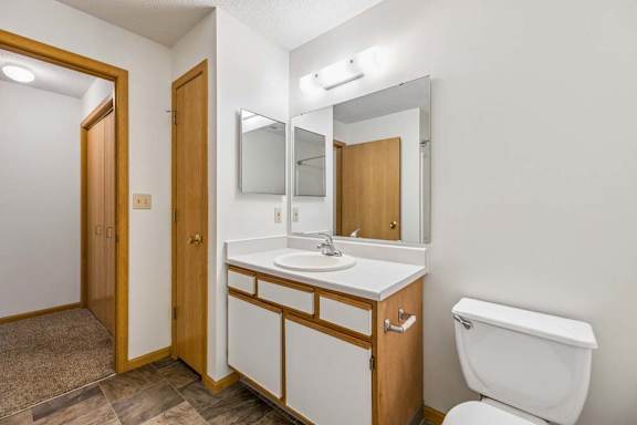 Apartment bathroom with a single-sink vanity, large wall mirror, toilet, and neutral finishes, with the hallway and bedroom visible through the open doorway.