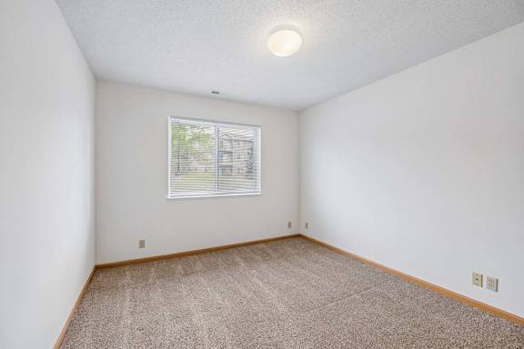 Bright, empty apartment bedroom with neutral carpet, white walls, wood trim, and a single window with blinds providing natural light.