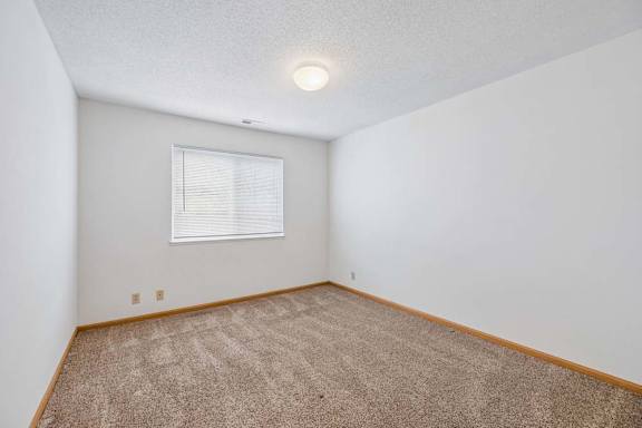 Bright, empty apartment bedroom with neutral carpet, white walls, wood trim, and a window with blinds that provides natural light.
