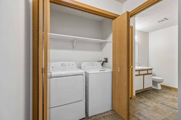 Apartment laundry closet with a full-size washer and dryer, bi-fold wood doors, and an adjacent bathroom featuring a single-sink vanity and tub/shower combination.