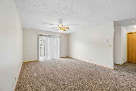 Spacious apartment living room with neutral carpet, a ceiling fan, vertical blinds over a sliding glass door, and an open layout leading to the hallway.
