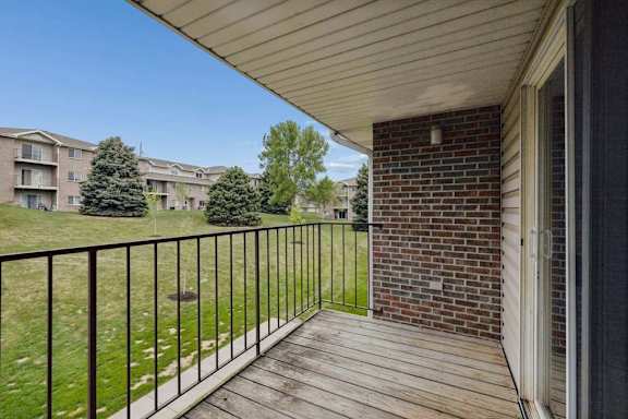 Covered apartment balcony with wood decking, metal railings, and views of landscaped green space and neighboring apartment buildings.