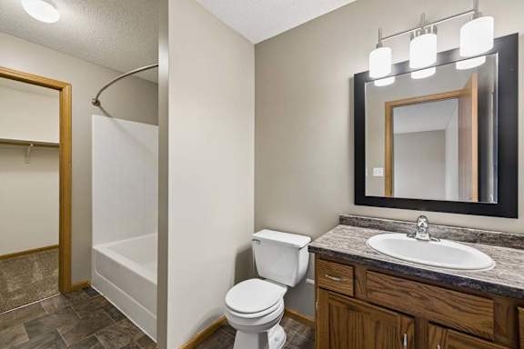 Apartment bathroom with a single-sink vanity, wood cabinetry, a granite-look countertop, a framed mirror with modern lighting, a tub-shower combination, and an adjacent walk-in closet.