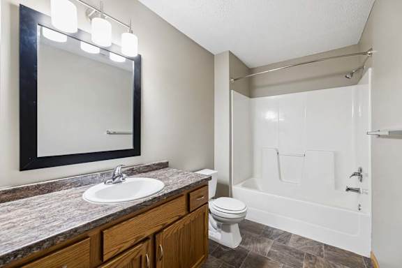 Apartment bathroom with a single-sink vanity, wood cabinetry, a granite-look countertop, a framed mirror with modern lighting, and a tub-shower combination with a white surround.