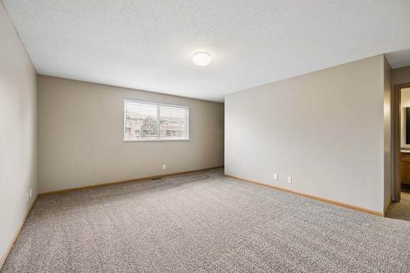 Spacious apartment bedroom with carpeted flooring, neutral walls, a large window providing natural light, and an open doorway leading to the adjoining bathroom.