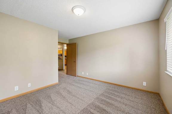 Bright apartment bedroom with carpeted flooring, neutral walls, a window with blinds providing natural light, and an open doorway leading toward the walk-in closet.