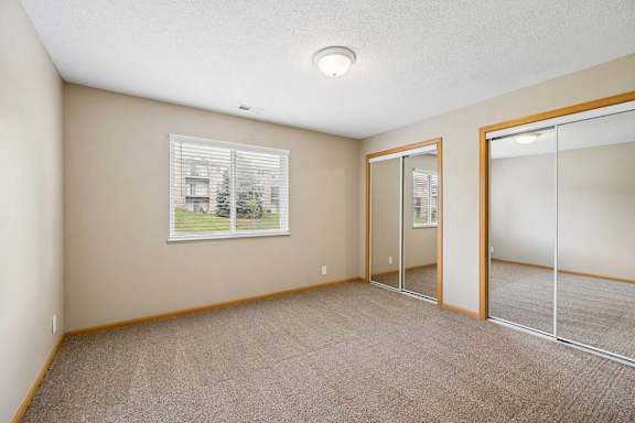 Bright apartment bedroom with carpeted flooring, a window providing natural light, and mirrored sliding closet doors for convenient storage.