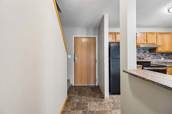 Apartment entryway with tile flooring, a wood front door, and a breakfast bar leading into the kitchen with wood cabinetry, black appliances, and a tile backsplash.