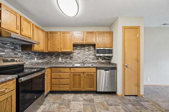 Apartment kitchen with wood cabinetry, laminate countertops, a tile backsplash, black and stainless steel appliances, and a functional U-shaped layout adjacent to the living area.