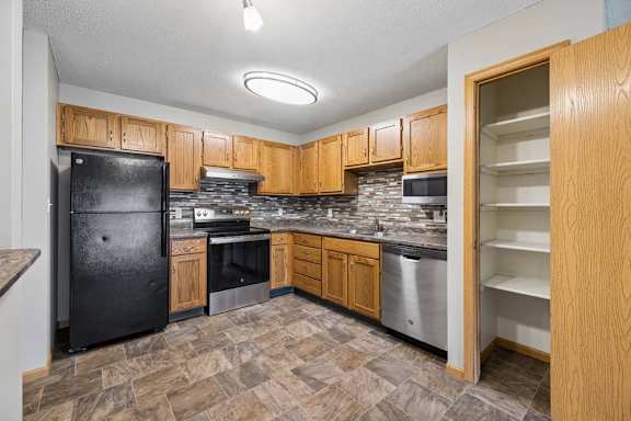Apartment kitchen with wood cabinetry, black appliances, a tile backsplash, laminate countertops, a pantry with built-in shelving, and a functional U-shaped layout.