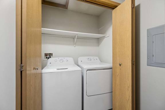 Apartment laundry closet with side-by-side washer and dryer, built-in shelf above, and wood bifold doors for convenient in-home laundry access.