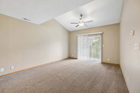 Spacious apartment living room with carpeted flooring, a vaulted ceiling, a ceiling fan, and a large sliding glass door with vertical blinds leading to a private patio or balcony.