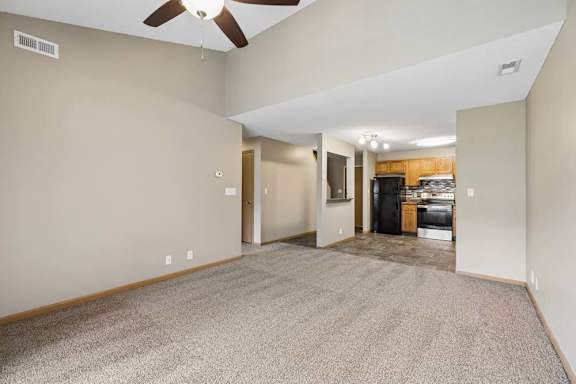 Open-concept apartment living room with carpeted flooring, a ceiling fan, vaulted ceilings, and a view into the kitchen featuring wood cabinetry and black appliances.